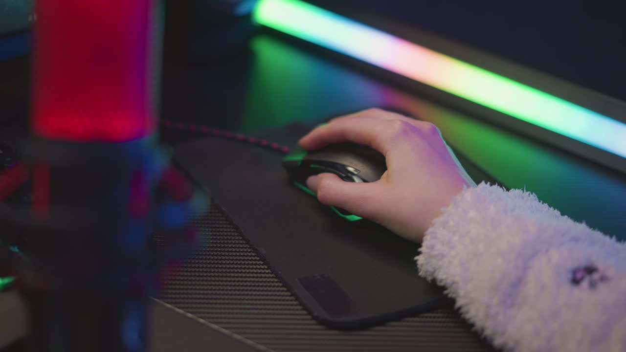 Close up of woman hand with polished nail in fluffy sweater using wired gaming mouse on black mousepad, surrounded by colorful ambient lighting from LED strip and microphone