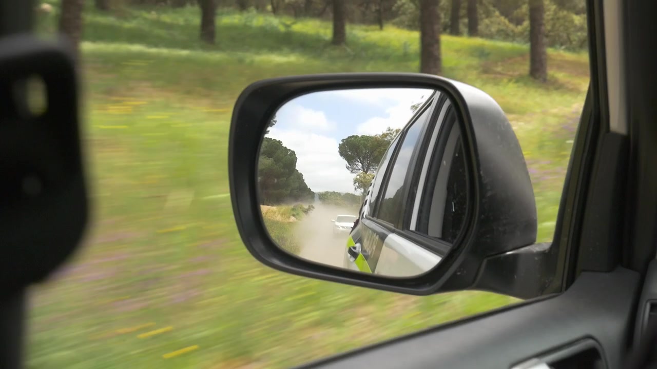 Specchietto retrovisore laterale di un veicolo della polizia (Unidad de Policía Adscrita a Andalucía) che pattuglia un'area forestale. Un'altra auto bianca segue sollevando polvere. Concetto di sicurezza e pattugliamento rurale