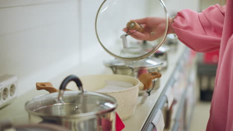 mano de una compradora inspeccionando una olla en una tienda de utensilios de cocina, la compradora está levantando cuidadosamente la tapa de una olla de utensilio de cocina moderna con un mango de madera, rodeada de otras ollas en exhibición