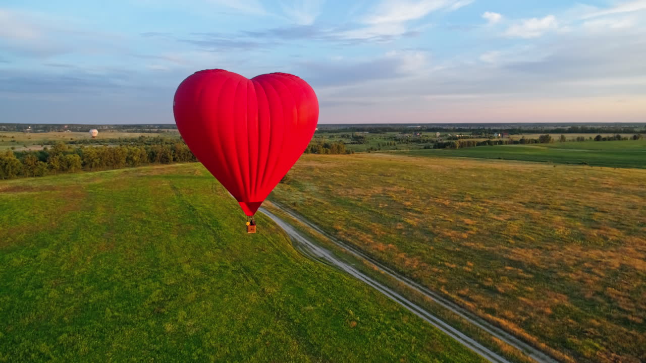 Hot air balloon flying over field. Romantic red aerostat in the heart shape moving slowly in the air. Fantastic view on the airship in summer.