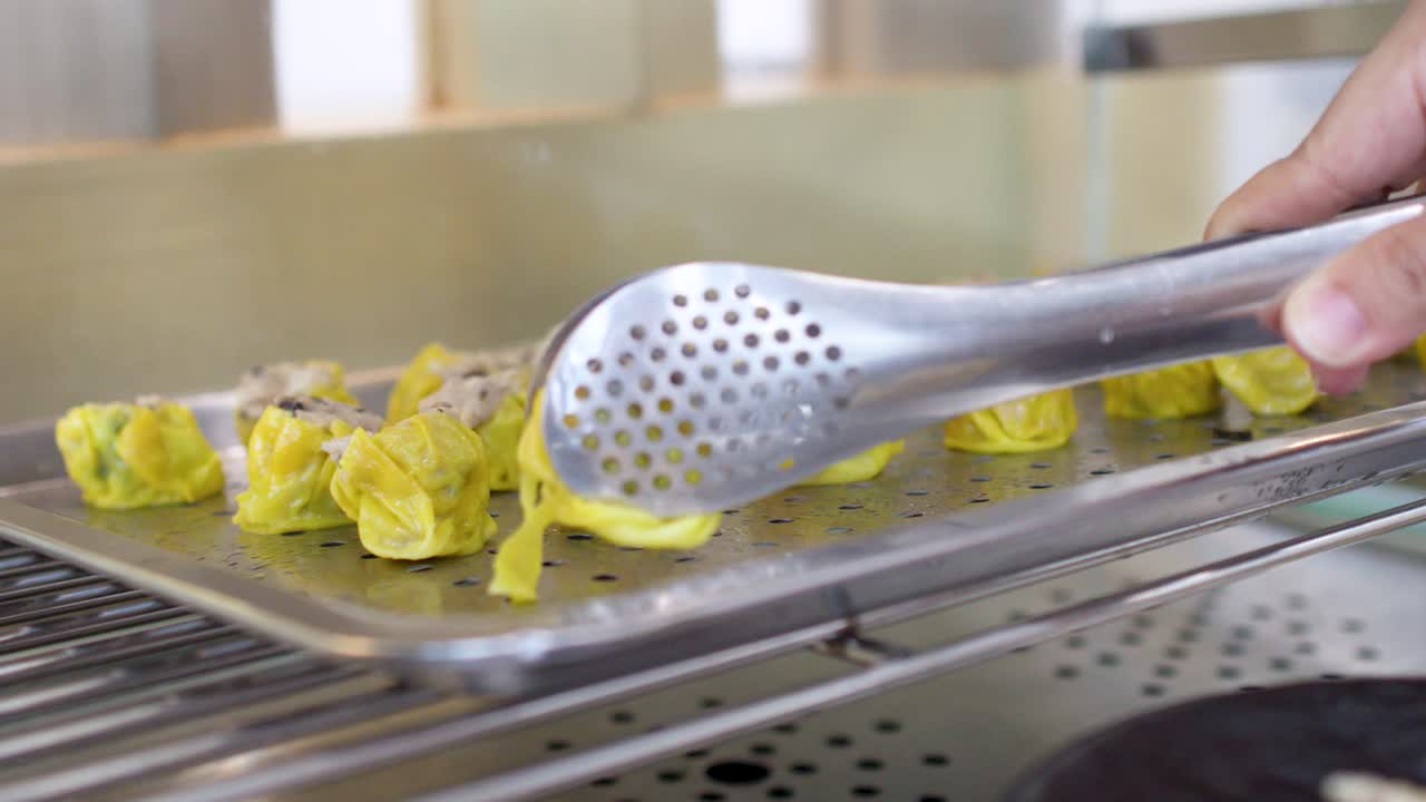 Metalltänger plockar upp gula siu mai-dumplings från en ångkorg på en restaurang.
