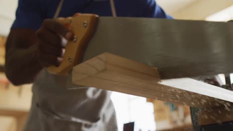 african american male carpenter cutting a wooden plank using a hand saw in a carpentry shop