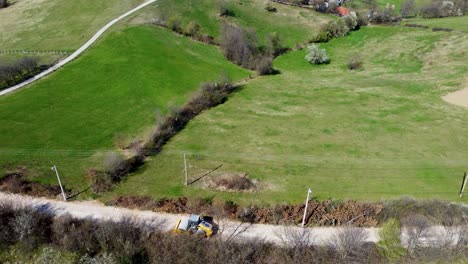 Yellow-single-smooth-drum-ride-on-vibratory-roller-compactor-moves-along-a-rocky-road-through-the-village-where-it-builds-infrastructure,-meadows-and-electric-poles-in-the-background,-drone-shot