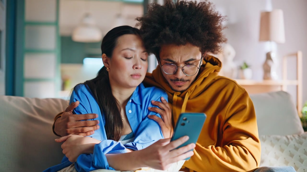 Multiethnic couple shocked by phone news at home. Boyfriend comforts girlfriend.
