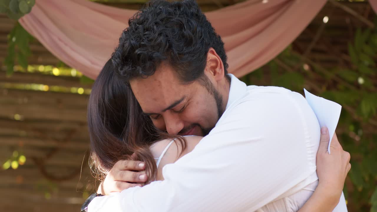 Coppia romantica che si abbraccia sotto un pergolato rosa, l'uomo con una maglietta bianca tiene in mano un biglietto piegato dopo averlo letto.
