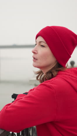 mujer con capucha roja y leggings negros descansa las manos en la barandilla decorativa de metal por un río tranquilo durante el invierno, suelo cubierto de nieve, puente lejano con vehículos en movimiento, atmósfera brumosa