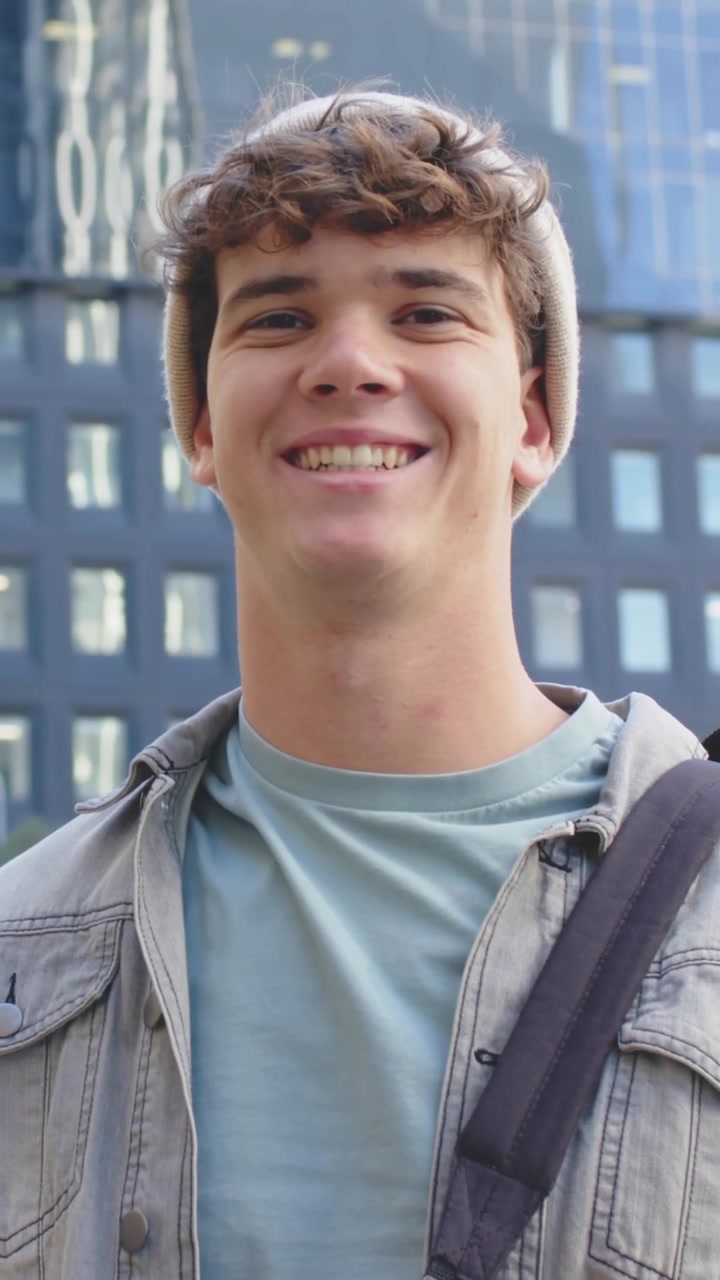 Video verticale: Un uomo sorridente con berretto inclina la testa e allarga il sorriso vicino a una facciata di vetro, mentre la telecamera zooma.
