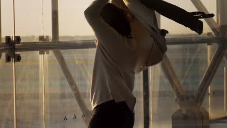 Young-Caucasian-woman-happy,-smiling,-and-twirling-alone-in-circle-with-her-winter-coat-above-her-head-inside-airport-on-sunny-day,-handheld-close-up-slow-motion