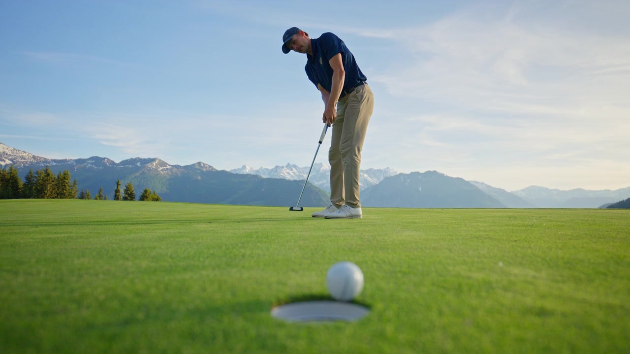 Inquadratura cinematografica dal basso di un golfista che imbuca un putt su un campo da golf di montagna a Verbier, Svizzera. La pallina da golf rotola sul green e cade nella buca con un drammatico scenario alpino sullo sfondo