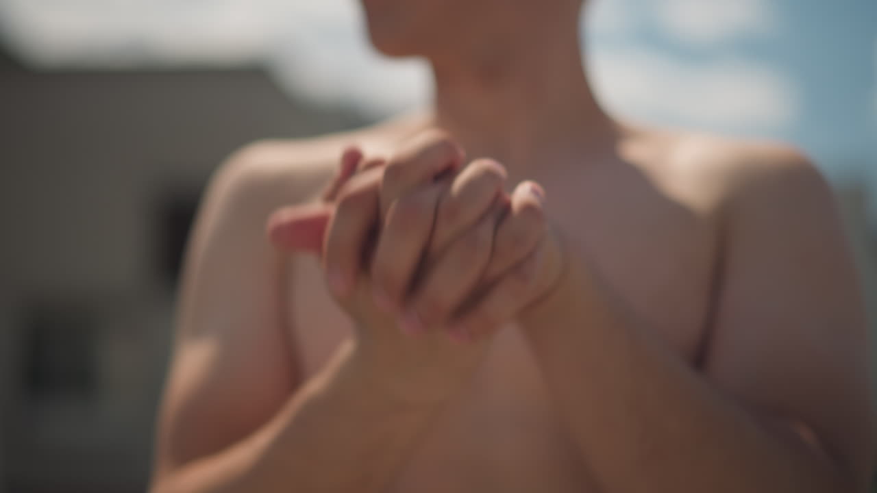 Мужчина с голым торсом сложил руки на крыше, крупный план переплетенных пальцев и запястий в лучах солнца, размытый городской пейзаж на заднем плане, медитативная разминка перед тренировкой, мягкое боке и теплые тона