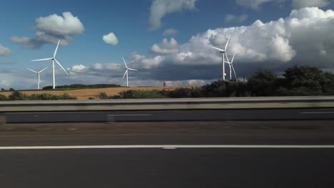 Wind-farm-from-seen-from-a-road-with-burning-fossil-fuel-gas-flaring-from-a-chimney-in-the-distance-along-side-green-fields