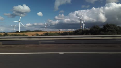 wind farm from seen from a road with burning fossil fuel gas flaring from a chimney in the distance along side green fields