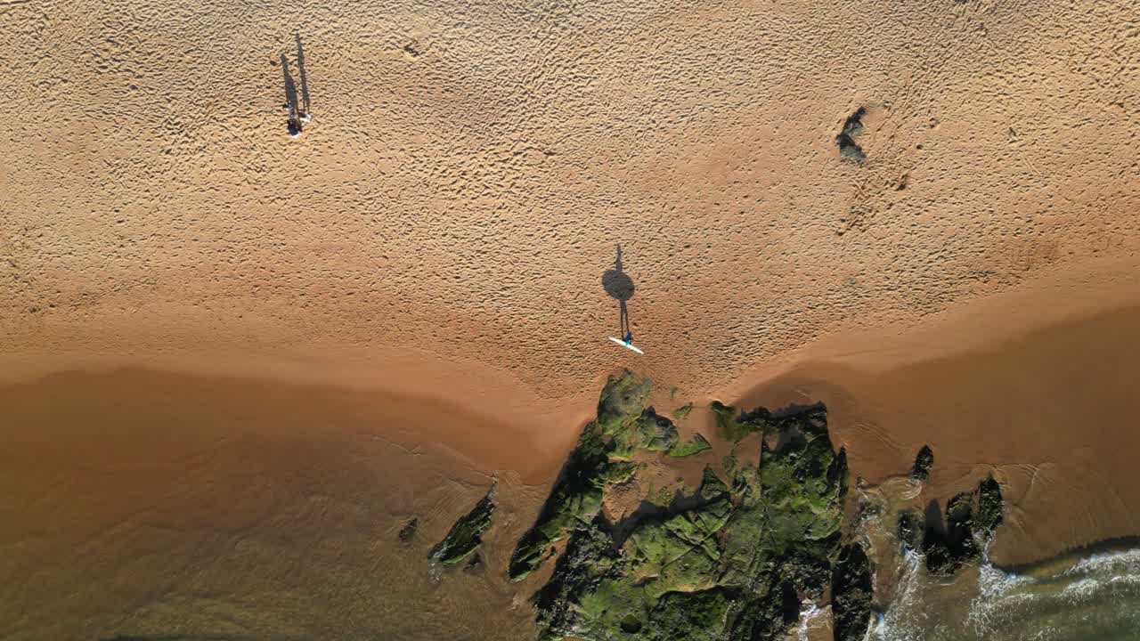 Seen from above a surfer walking along the seashore carrying his surfboard, with his shadow reflecting on the sand.Portugal