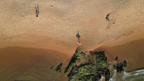 seen from above a surfer walking along the seashore carrying his surfboard, with his shadow reflecting on the sand.portugal