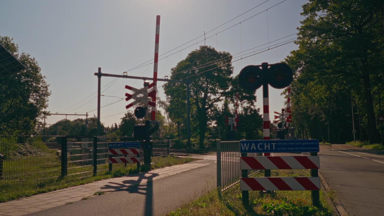 Static view of a railway crossing in the Netherlands with red and white barrier gates, level crossing signs and railway tracks in a peaceful outdoor setting with blue skies.