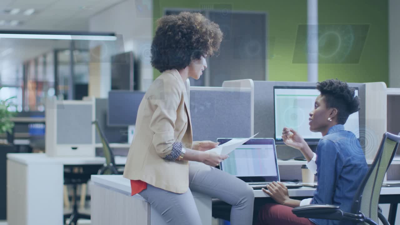 Perched woman sliding charts at laptop, seated peer typing and analyzing business data with HUD