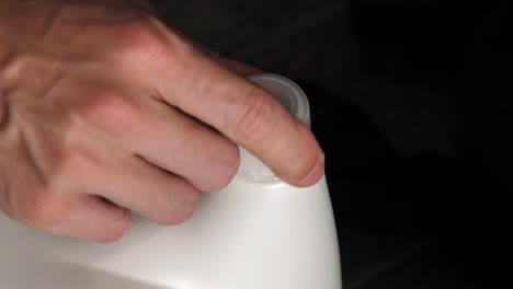 mano masculina atornillando la tapa superior de una botella de lavandería de plástico blanco con fondo oscuro, tarea doméstica