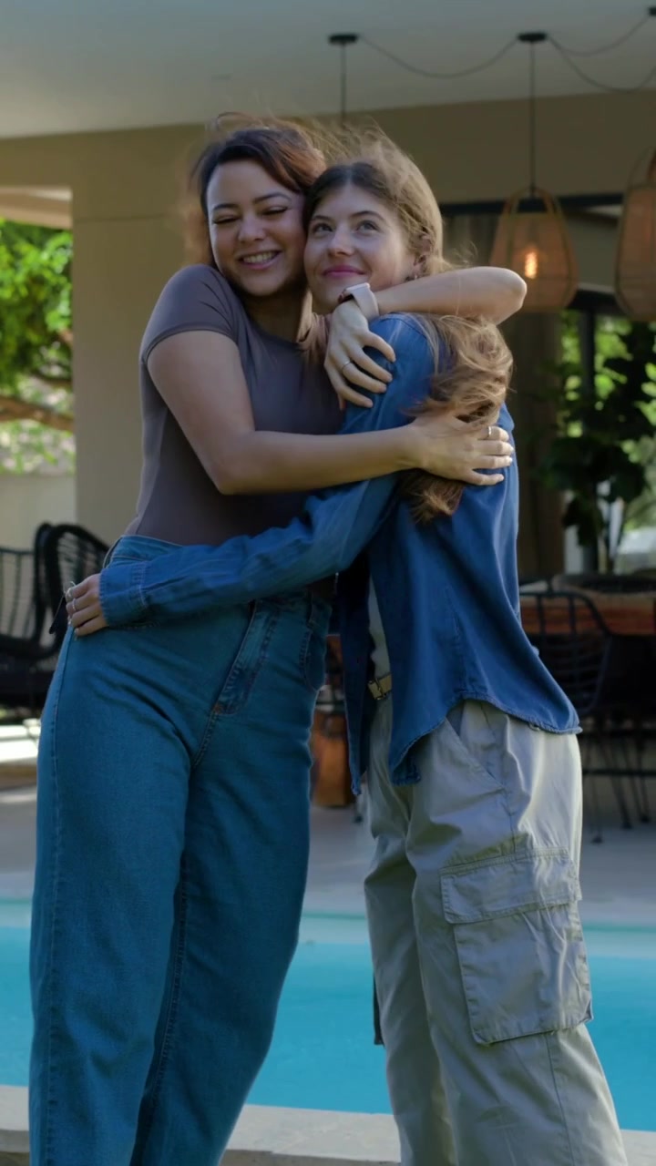 Video verticale: Donna a sinistra che ride, condivide una bottiglia rossa con un'amica in piscina, indossa una camicia di jeans e un top marrone.