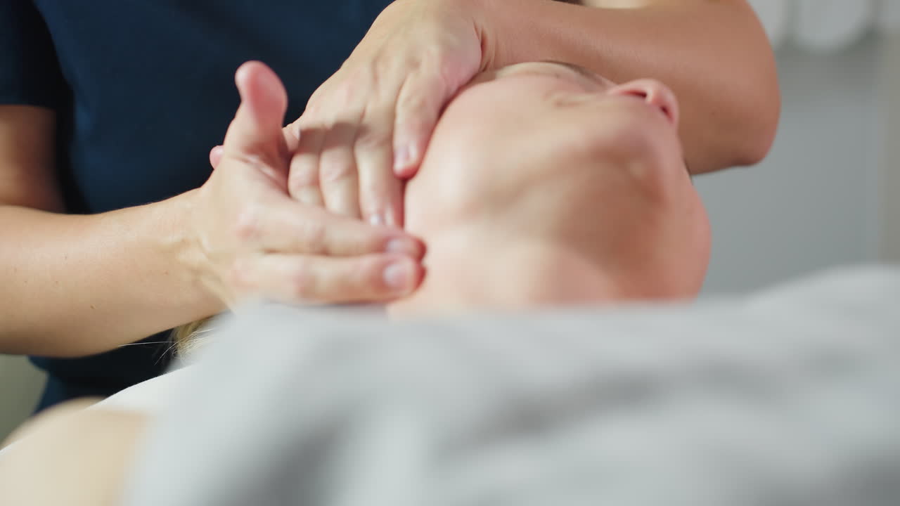 Wellness technician massaging patient neck gently in bright spa room with white background during relaxing facial therapy session showcasing expert hands and serene tranquil environment