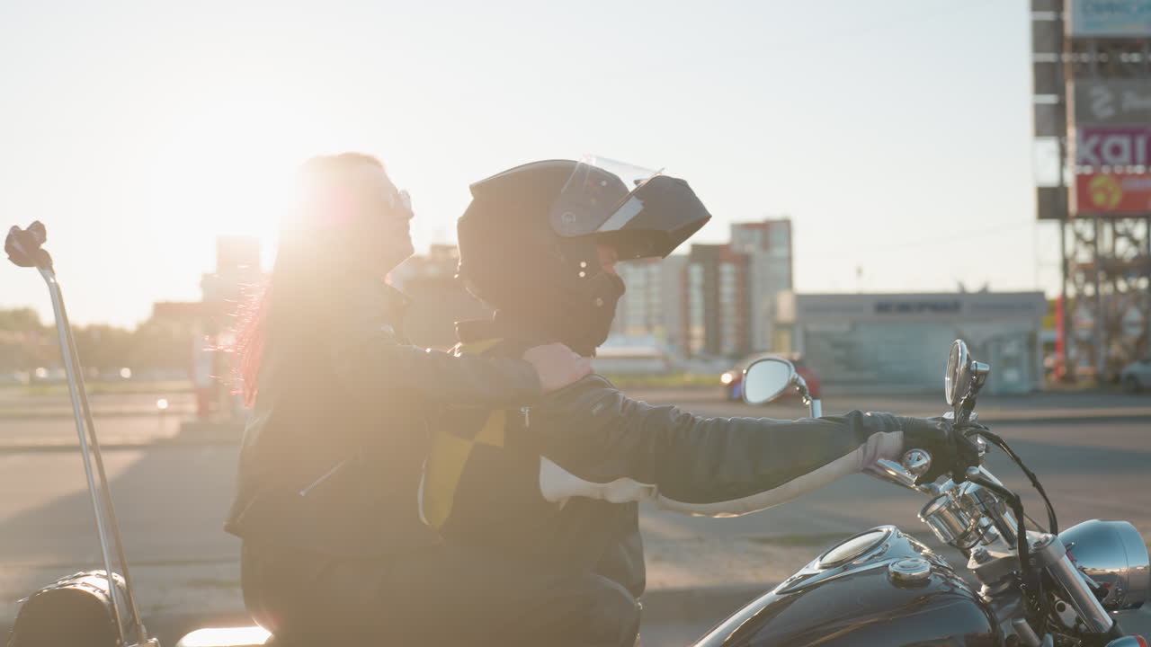 Woman in leather climbs onto motorcycle behind rider seated in sunlight, legs stepping on footrest while holding balance, warm glow highlighting chrome details and blurred city background