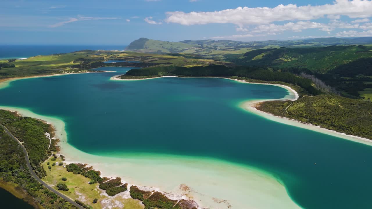 Straight aerial flyover of Kai Iwi Lakes revealing turquoise water, white beaches, and rolling farmland with ocean beyond New Zealand