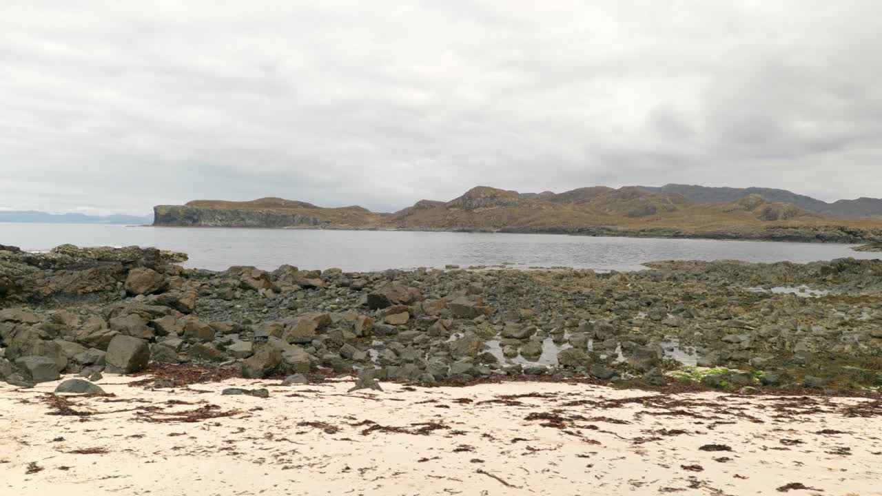 천천히 옆으로 움직이며 스코틀랜드의 모래사장을 따라 패닝 샷을 배경으로 바위, 바다, 산을 바라보고 있습니다.