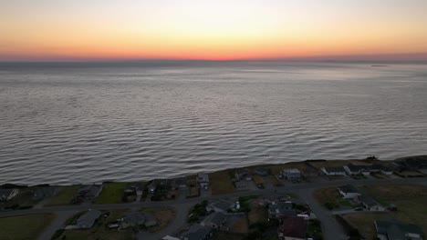 Vista-Aérea-Alejándose-Del-Brillo-Anaranjado-De-La-Puesta-De-Sol-Para-Revelar-Una-Comunidad-Frente-Al-Mar