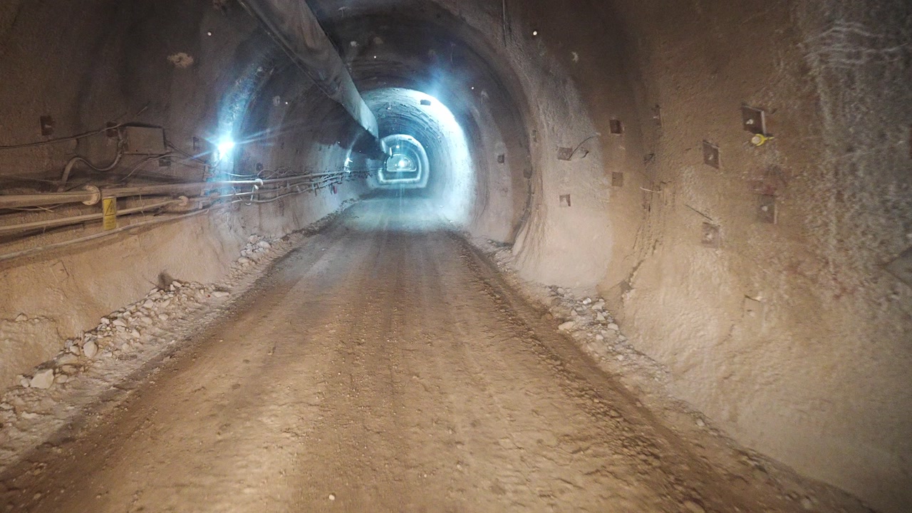 A first-person view from inside a vehicle shows a hand on the steering wheel as a dusty windshield frames a construction site outside, conveying focus, travel, and rugged work.