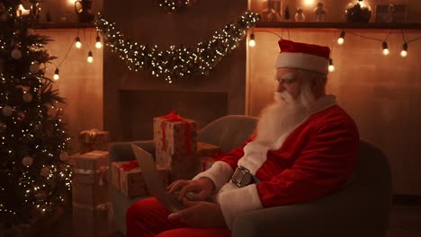 santa claus sits next to a christmas tree on a comfortable soft sofa, holds a laptop in his lap and answers emails from young children.