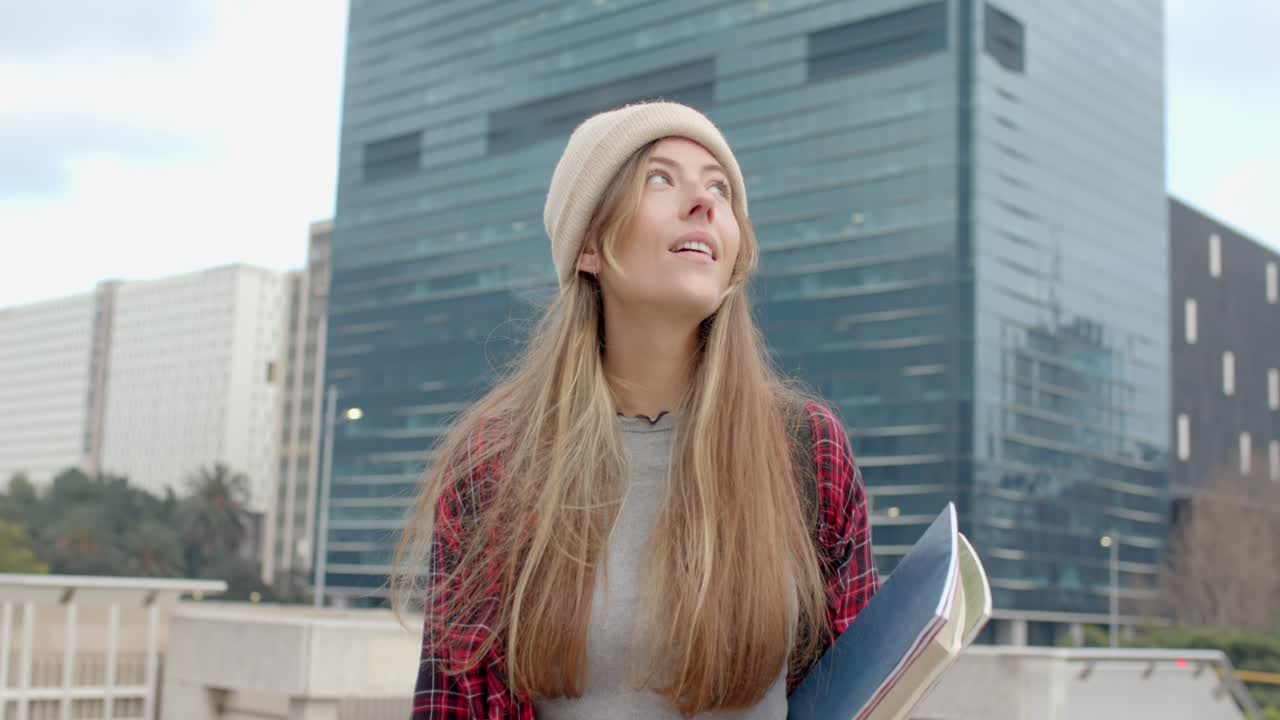Arriving on terrace with notebooks folders, woman in twenties gazing upward and smiling at building