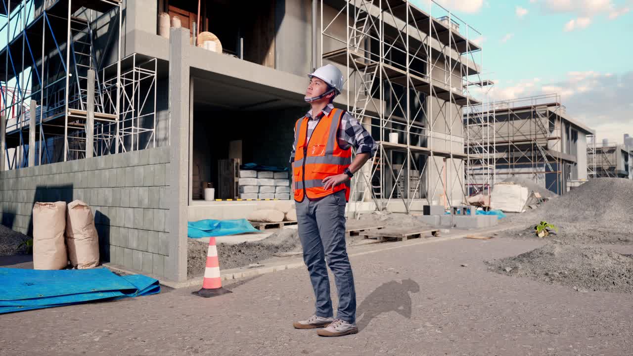 Full Body Side View Of Asian Male Engineer Wearing Safety Helmet Looking Around While Standing With Arms Akimbo At Building & Construction Site in Progress to New House