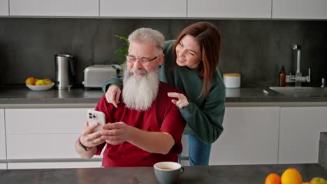 A-happy-and-cheerful-elderly-man-with-gray-hair-and-a-lush-beard,-together-with-his-adult-brunette-daughter-in-a-green-jacket-take-a-selfie-using-a-white-smartphone-in-a-modern-kitchen