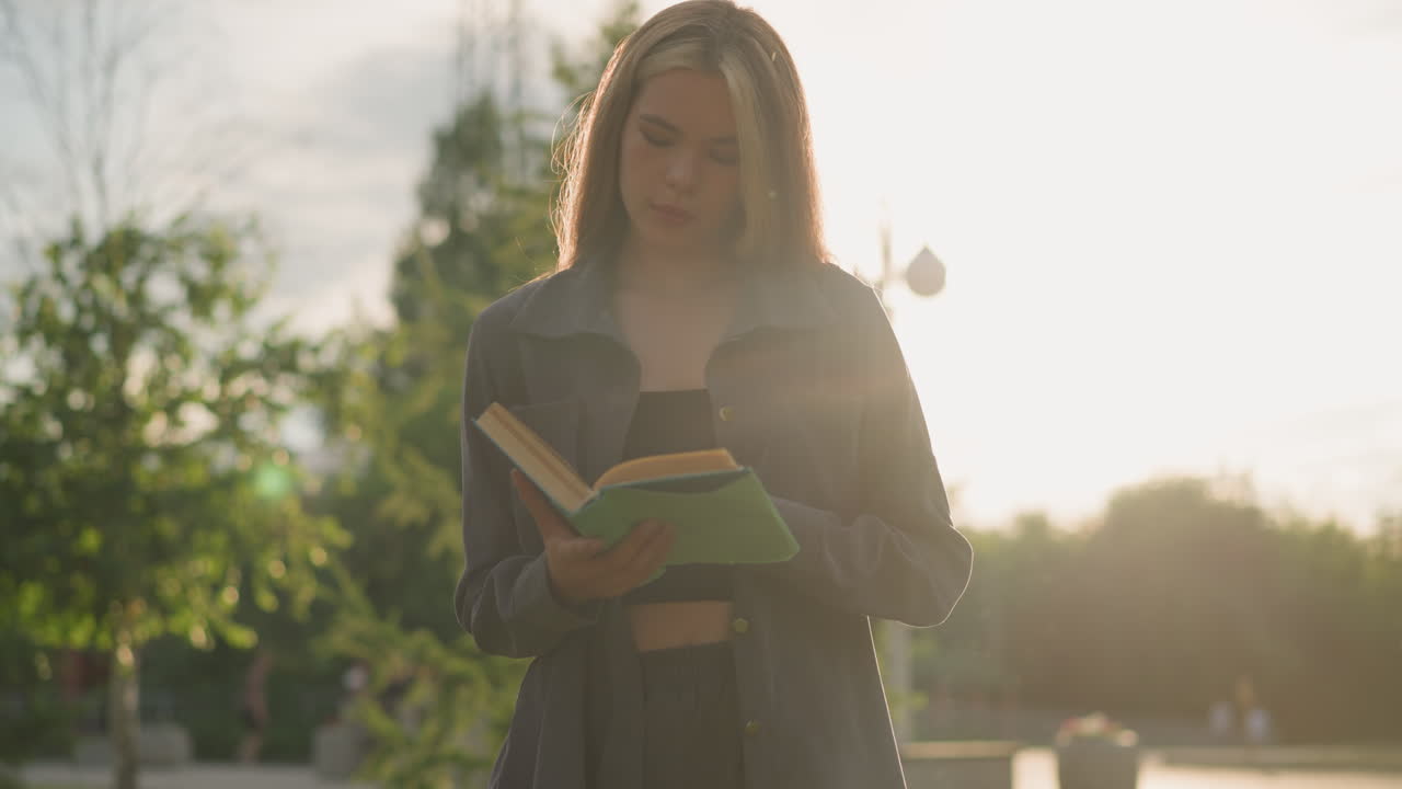 회색 옷을 입은 여인이 야외에 서서 머리를 약간 기울여 책 페이지를 뒤집고, 빛이 그녀의 배경으로 부드럽게 반사되어, 흐릿한 나무와 건물을 특징으로합니다.