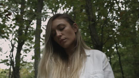 Orbit-low-angle-shot-of-woman-with-long-blonde-hair-looking-at-trees-in-a-forest