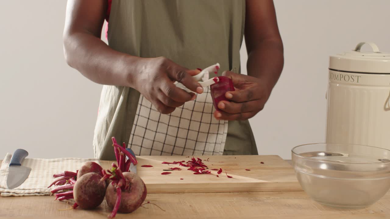 Uomo afroamericano di mezza età che pela barbabietole con un pelapatate a Y sul bancone di casa, con un grembiule color oliva, per il compost.