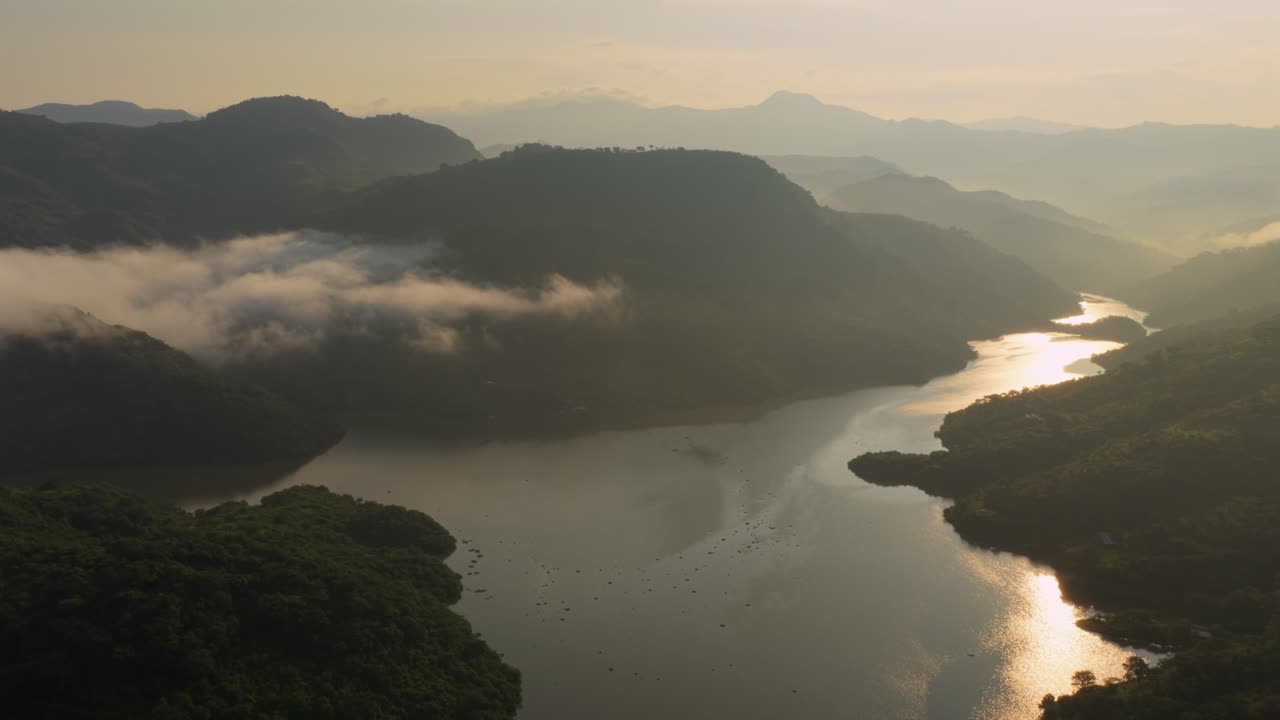 Drone video capturing serene Presa El Carrizo, Tamazula de Gordiano at sunrise. Calm waters reflect the rising sun, surrounded by lush hills and mist, creating a tranquil scene