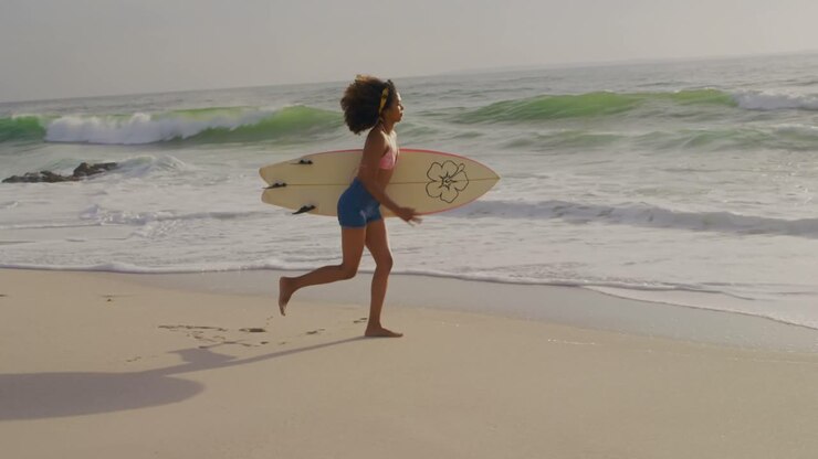 Seitenansicht einer afroamerikanischen Surferin, die mit Surfbrett am Strand rennt 4K
