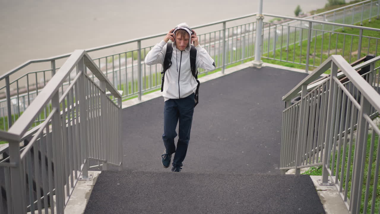후드티를 입고 백팩을 멘 흑인 남성이 계단을 올라가면서 휴대폰을 확인하고, 헤드폰을 조절하며, 흐린 하늘 아래 금속 난간과 아스팔트 보도가 있는 착륙 지점으로 올라갑니다. 배경에는 강이 살짝 보입니다.