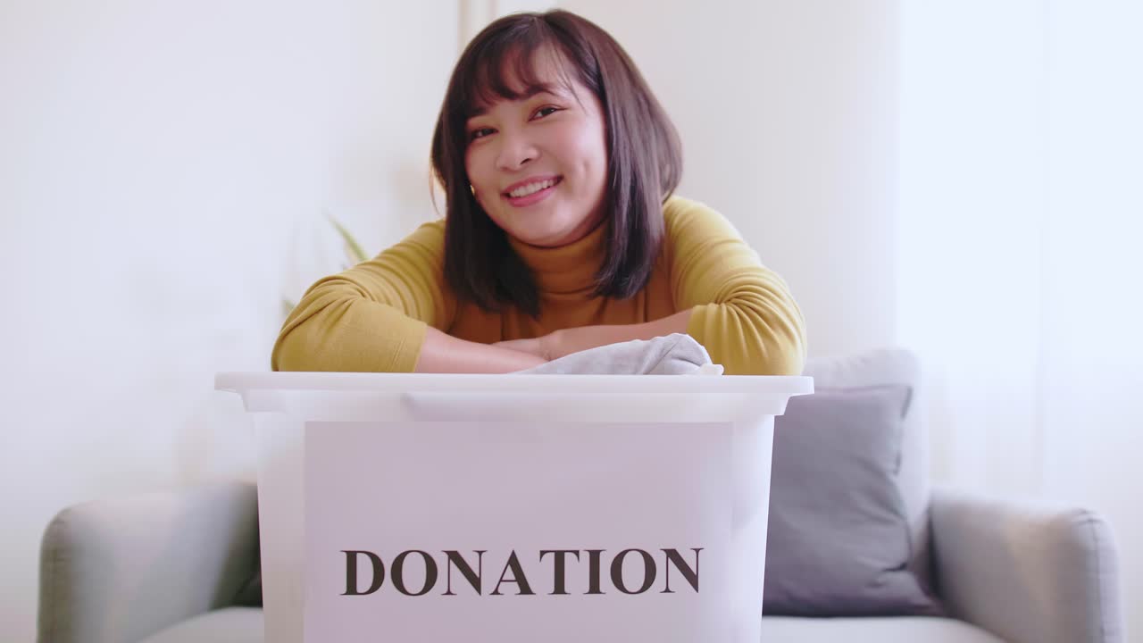 Portrait of young Asian woman looking at camera holding box with clothes for donation