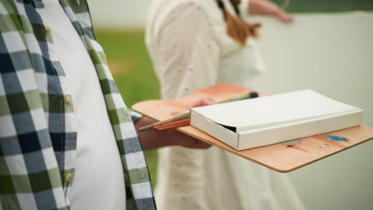 A close-up shot of a person holding a wooden palette with paintbrushes and a white paint box, preparing to paint outdoors. The individual wears a white t-shirt with a green and blue checkered flannel