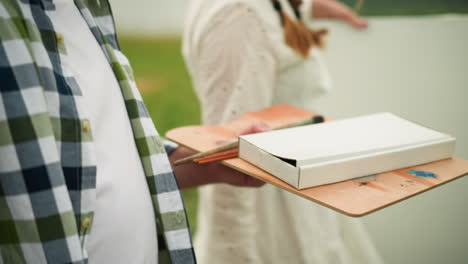a close-up shot of a person holding a wooden palette with paintbrushes and a white paint box, preparing to paint outdoors. the individual wears a white t-shirt with a green and blue checkered flannel