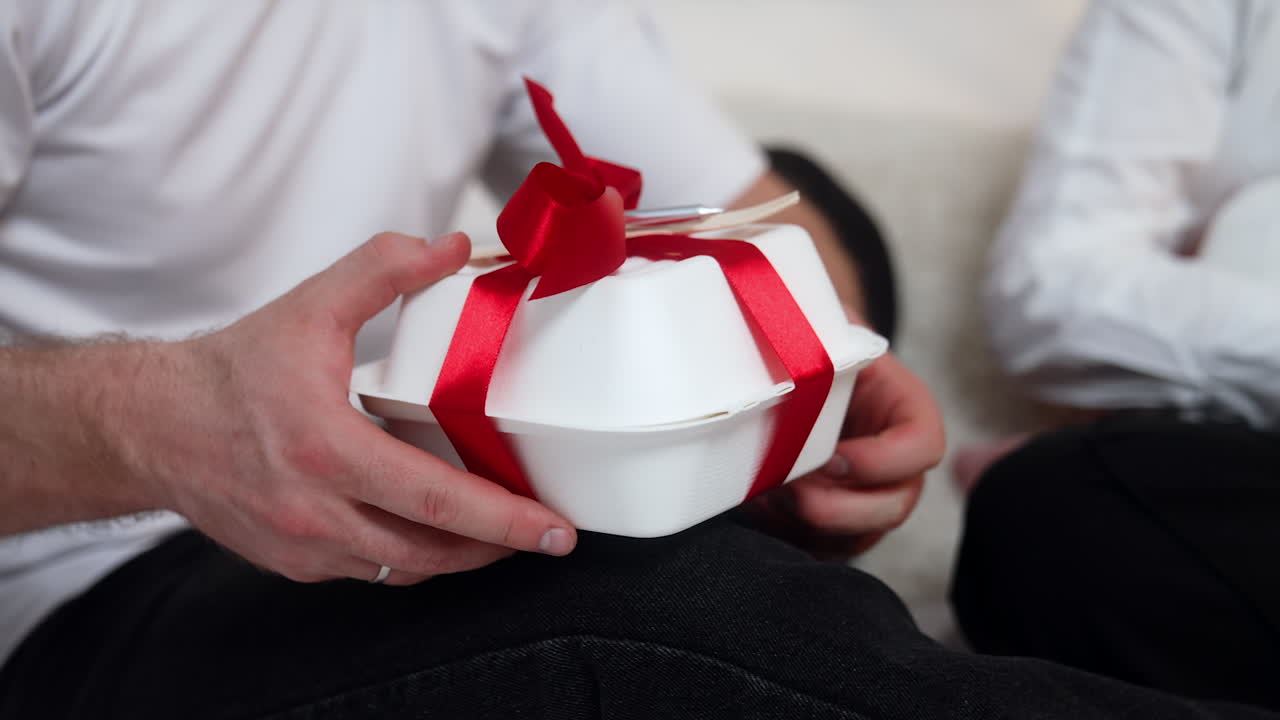 Food box with red ribbon and fork. Male hand take the box and pulls the ribbon to untie it. Close up.