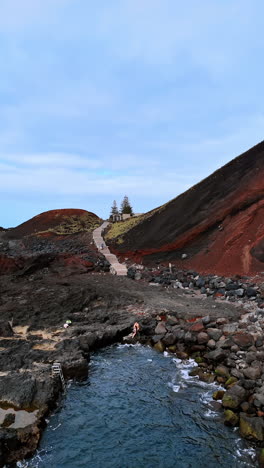 lonely house with two pine trees on the rocky shore near the mountain. drone rising above the waterscape. azorean islands. vertical video.