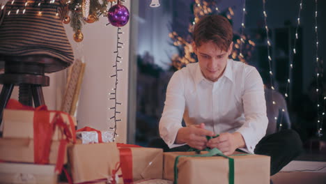 man tying ribbon on wrapped gift box at home during christmas