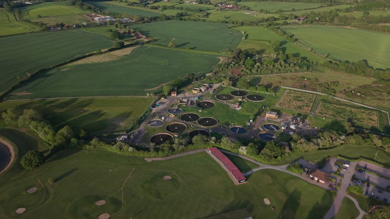 An expansive aerial view captured during golden hour reveals a water treatment plant with multiple circular tanks situated amidst lush green countryside.