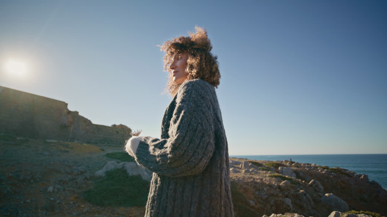Autumn lady standing ocean cliff at sunshine back view. Model holding dry flower