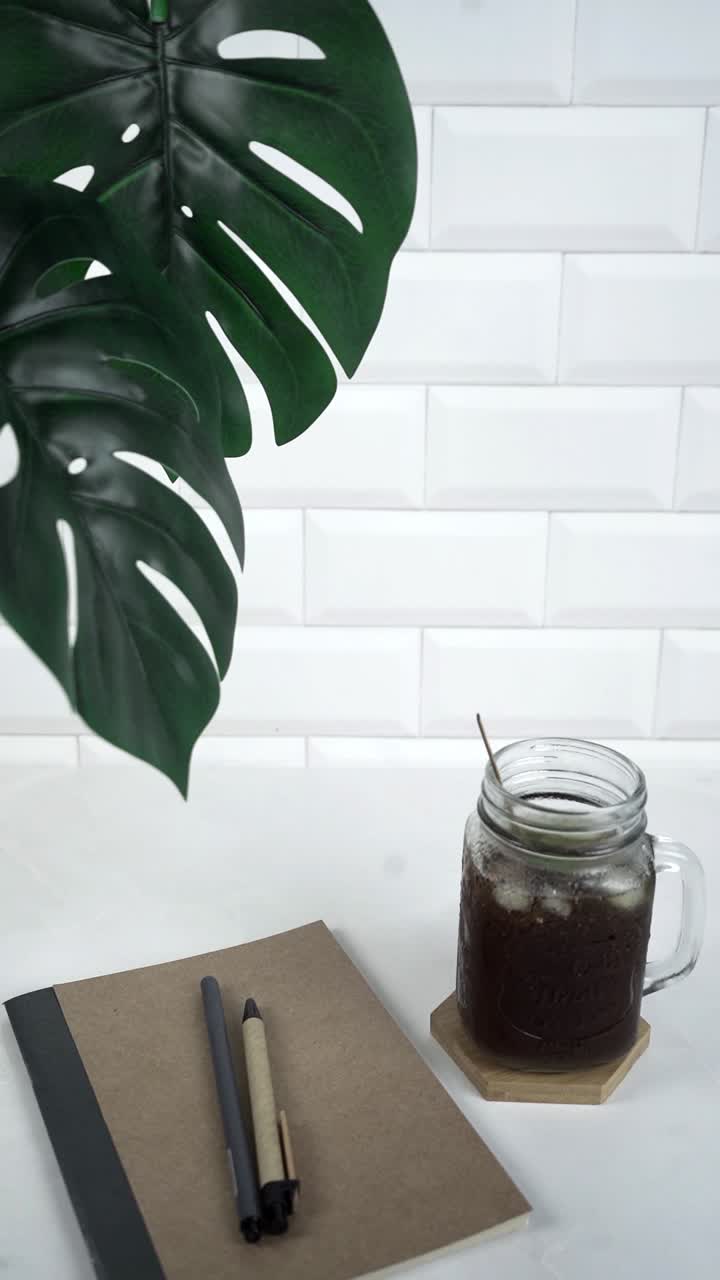 Foglia di Monstera, caffè freddo con ghiaccio, notebook e penne su scrivania bianca