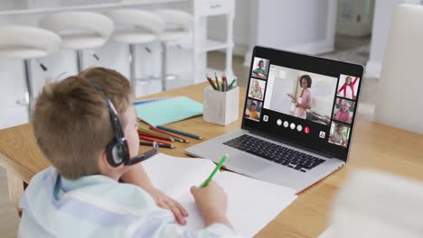 Schoolboy-using-laptop-for-online-lesson-at-home,-with-diverse-teacher-and-class-on-screen