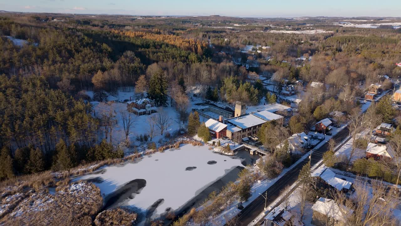 Winter drone ascent over a frozen pond and forested rural community, revealing the historic mill complex surrounded by snow-covered homes and peaceful countryside
