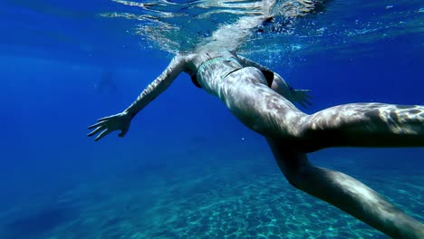 woman in a bikini is swimming underwater in a crystal-clear blue ocean with the sunlight filtering through the surface, creating shimmering patterns on her skin and the sandy bottom